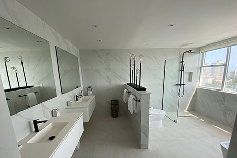 Modern bathroom with white marble walls and floors, dual floating vanities, and walk-in glass shower with black fixtures. 